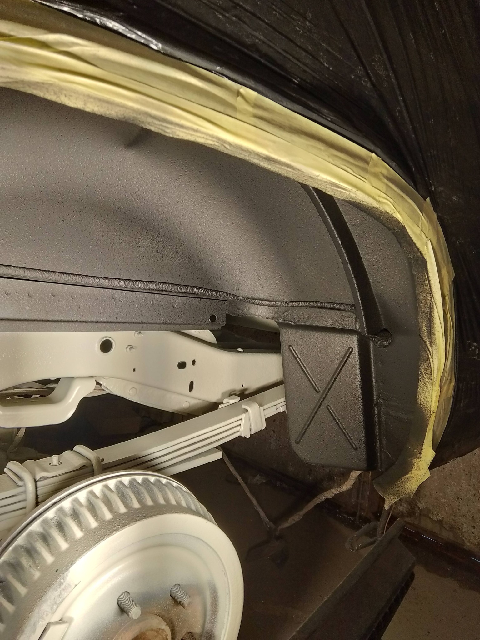 Antykorozyjne zabezpieczenie podwozia samochodu z użyciem taśmy maskującej, po nałożeniu czarnej powłoki ochronnej. Widoczne elementy zawieszenia i nadkola.