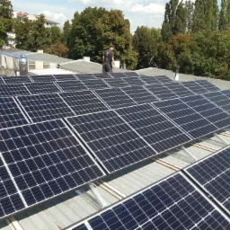 Instalacja paneli słonecznych na dachu budynku, widok z góry na rozległą powierzchnię paneli, z pracownikiem w oddali i drzewami w tle.