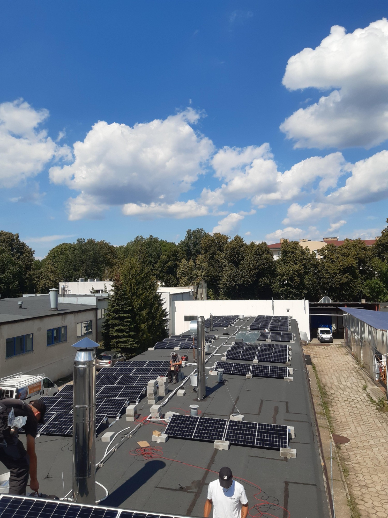 Montaż paneli słonecznych na płaskim dachu budynku przemysłowego, widoczne narzędzia i materiały instalacyjne, błękitne niebo z chmurami w tle.