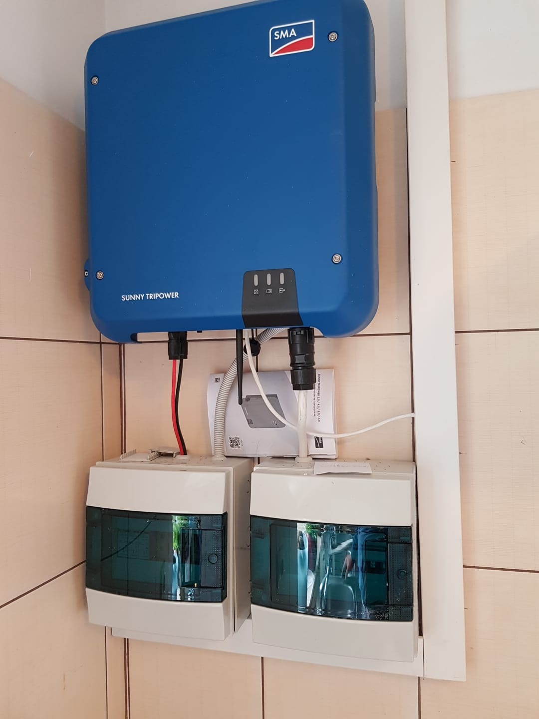Niebieski falownik SMA Sunny Tripower zamontowany na ścianie z dwoma skrzynkami rozdzielczymi poniżej, widoczne okablowanie i dokumentacja techniczna.