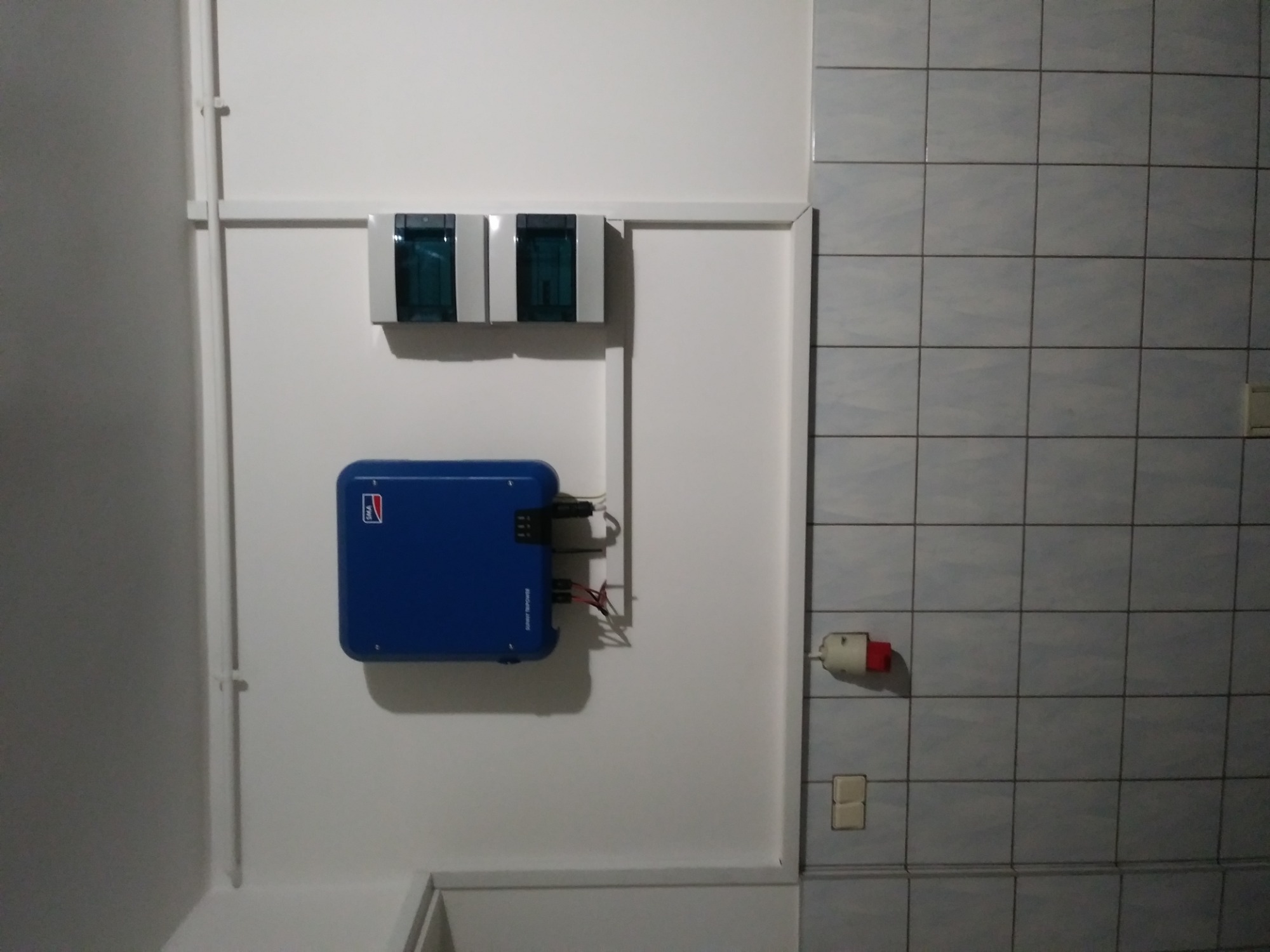 Niebieska skrzynka inwertera z podłączonymi kablami, zamontowana na białej ścianie w pomieszczeniu z częściowo wyłożoną kafelkami ścianą, obok widoczne dwie małe skrzynki elektryczne.