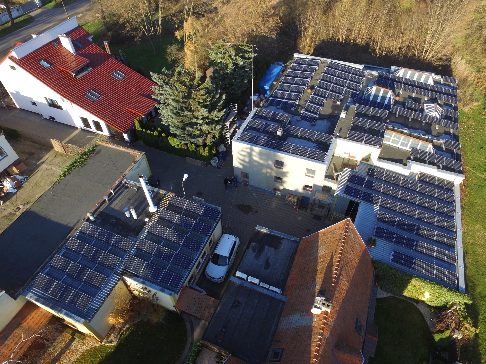Widok z góry na budynki z instalacją paneli słonecznych na dachach, biały samochód zaparkowany na dziedzińcu, drzewa iglaste i liściaste w tle.
