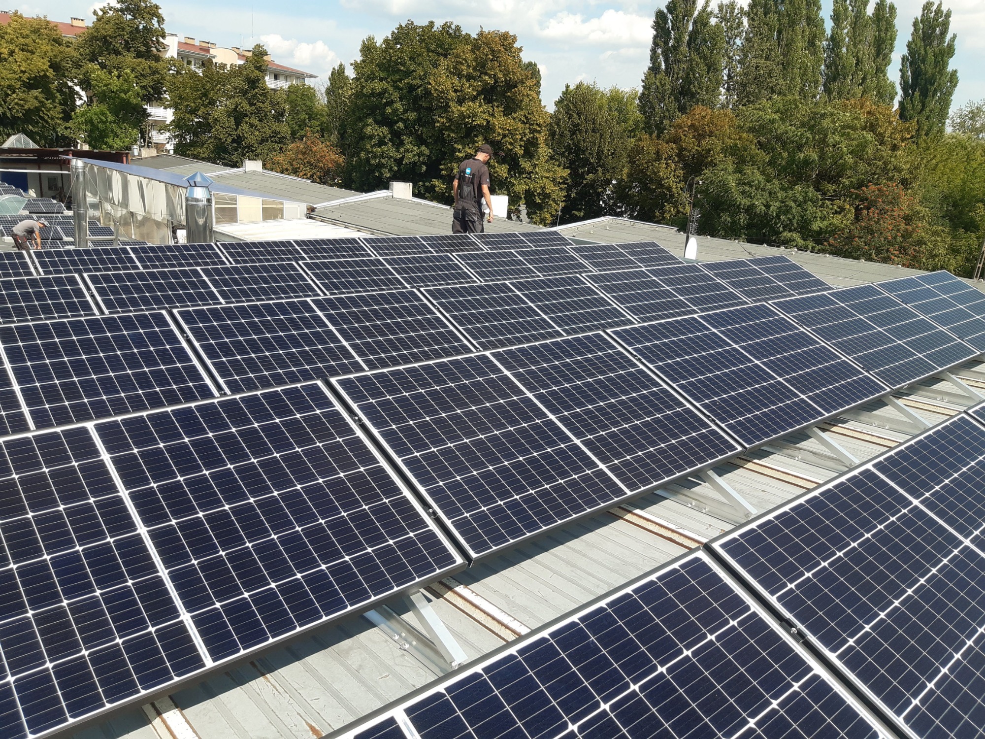 Instalacja paneli słonecznych na dachu budynku, widok z góry na rozległą powierzchnię paneli, z pracownikiem w oddali i drzewami w tle.