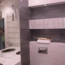 REMIS-AVS sp. z o. o. - Wnętrze łazienki w trakcie wykańczania w Warszawie, widoczne szare płytki, lustro z odbiciem grzejnika, zabudowa WC z silikonem na górze i niedokończona umywalka z wyciętymi otworami.