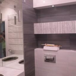 REMIS-AVS sp. z o. o. - Nowoczesna łazienka w trakcie wykańczania, widoczne szare płytki, podwieszana toaleta z przyciskiem spłukiwania, lustro z odbiciem grzejnika drabinkowego i osoby, tuba silikonu na zabudowie.