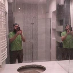 REMIS-AVS sp. z o. o. - Odbicie w lustrze łazienki w trakcie prac: mężczyzna robi zdjęcie otworu na umywalkę w blacie, widoczna instalacja pod umywalką, grzejnik drabinkowy i kabina prysznicowa w tle.