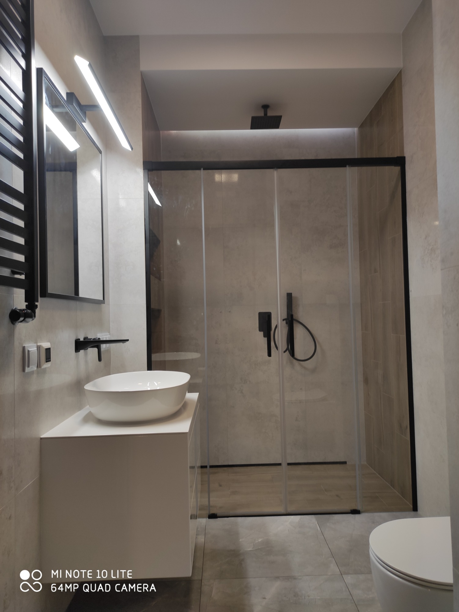 Nowoczesna łazienka z kabiną prysznicową typu walk-in z czarnymi ramami, owalną umywalką nablatową i czarną armaturą, oświetlona listwą LED nad lustrem.