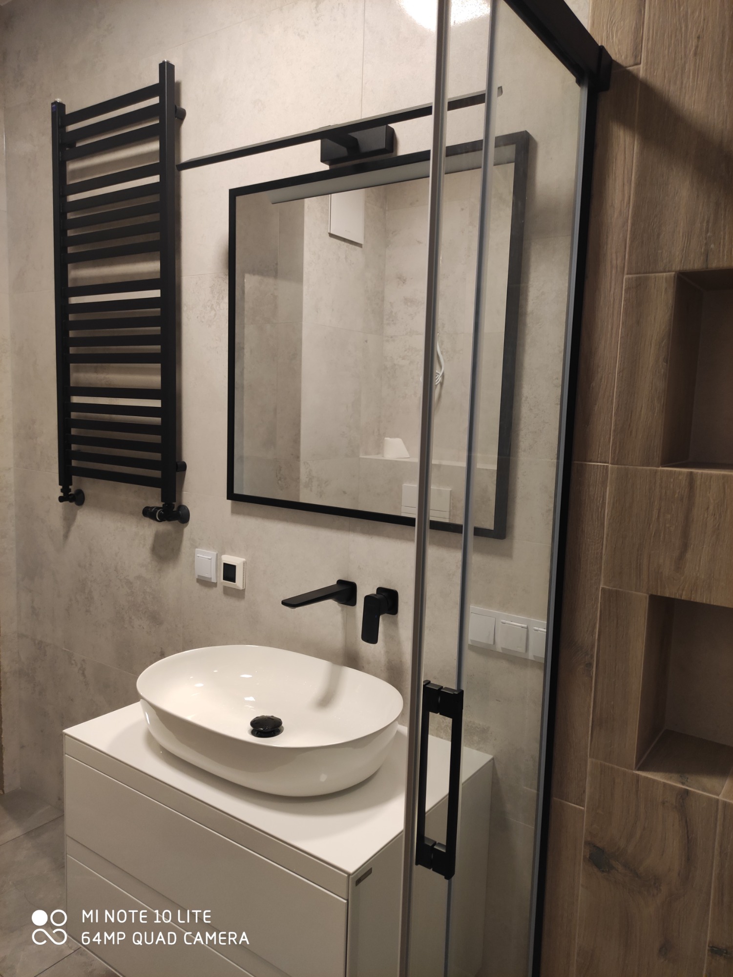 Nowoczesna łazienka z owalną umywalką nablatową, czarną armaturą, grzejnikiem drabinkowym i kabiną prysznicową z czarnymi profilami, wykończona płytkami w odcieniach szarości i drewna.