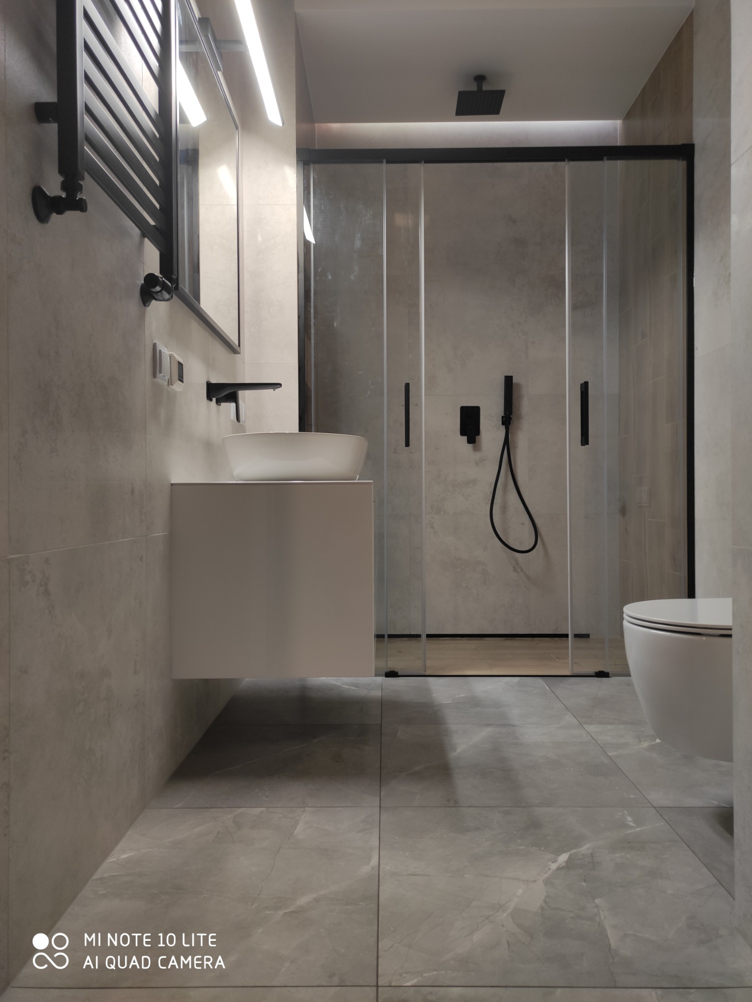 Nowoczesna łazienka z szarą glazurą, kabiną prysznicową typu walk-in z czarnymi ramami, umywalką nablatową na białej szafce i czarną armaturą, oświetleniem LED oraz grzejnikiem drabinkowym...