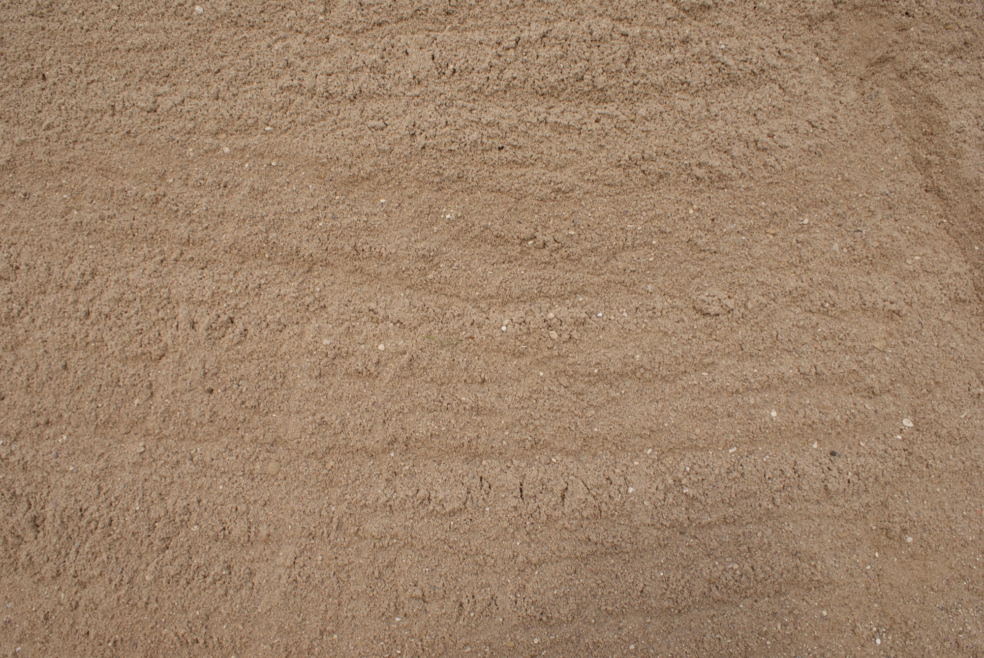 Beżowy, drobnoziarnisty materiał sypki z widocznymi śladami po przesuwaniu, prawdopodobnie piasek budowlany.