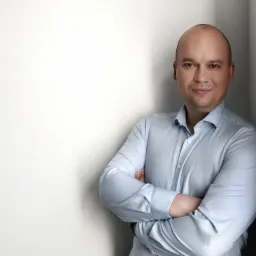 Portret uśmiechniętego mężczyzny w błękitnej koszuli stojącego pod ścianą z założonymi rękami.