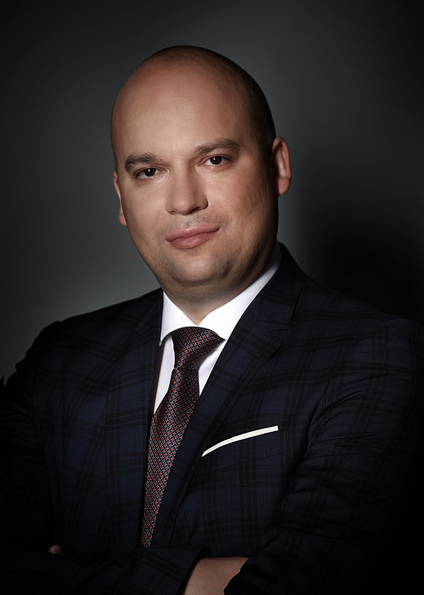 Portret mężczyzny w ciemnym garniturze w kratę i wzorzystym krawacie, z rękami skrzyżowanymi, na ciemnoszarym tle.