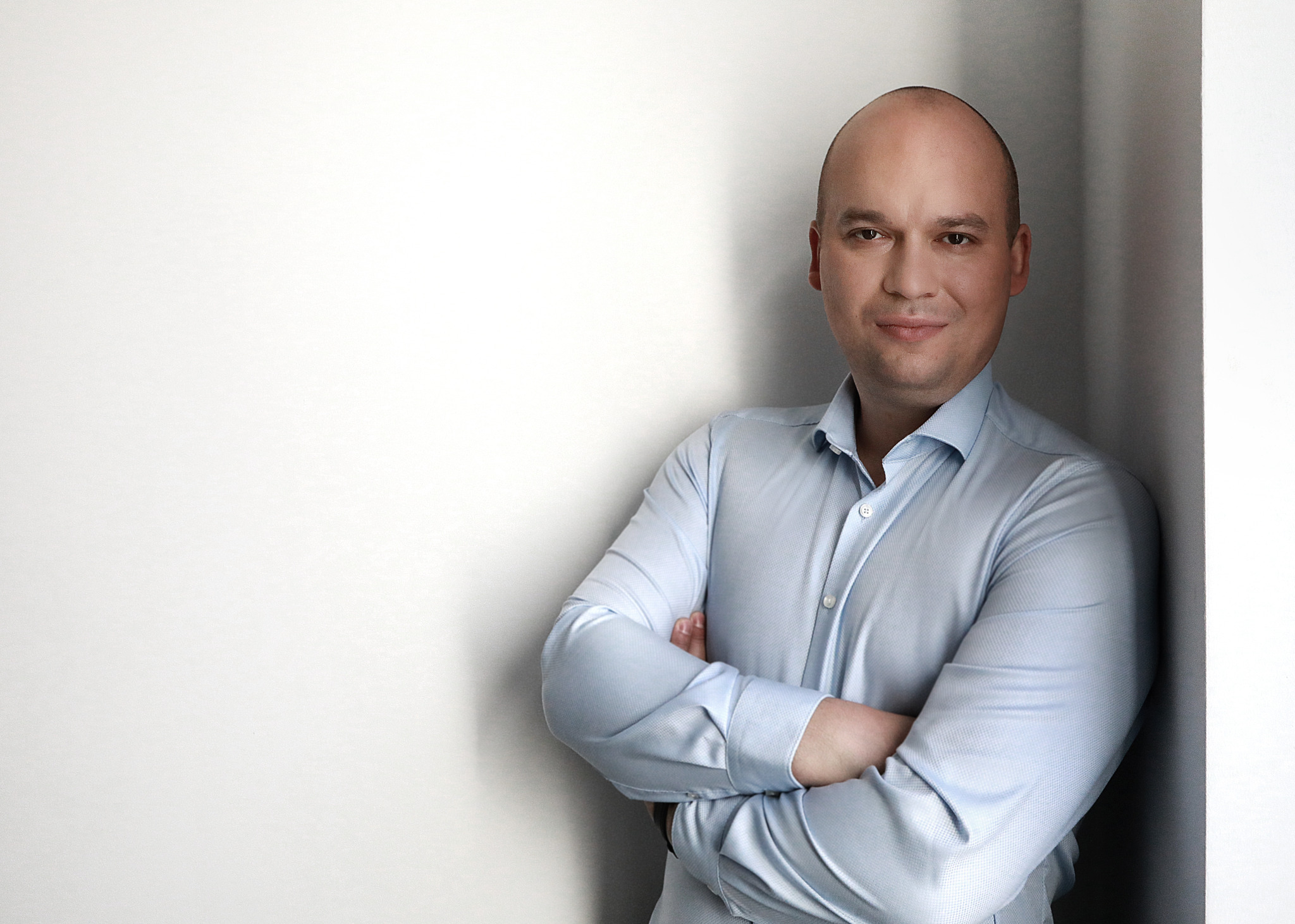 Portret uśmiechniętego mężczyzny w błękitnej koszuli stojącego pod ścianą z założonymi rękami.