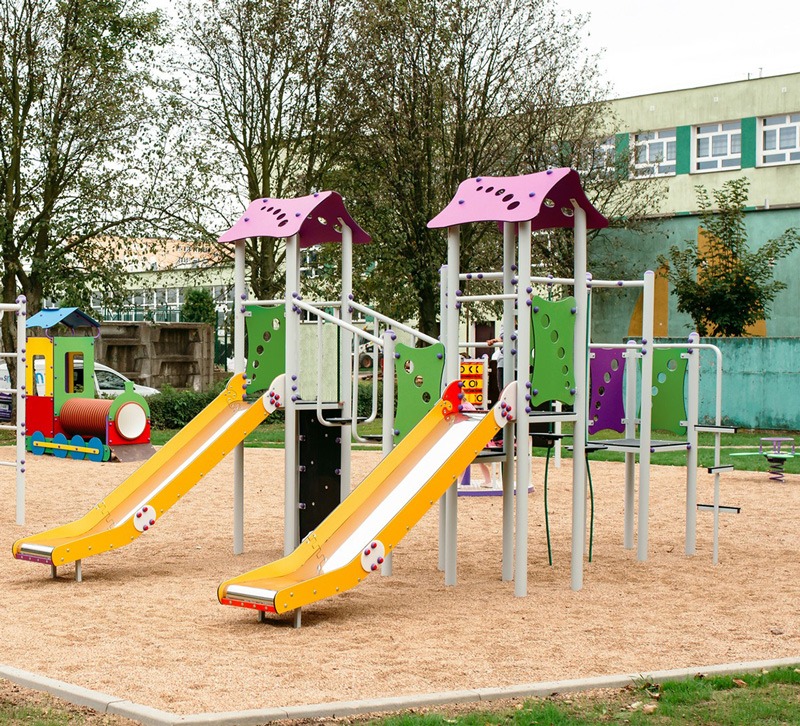 Kolorowy plac zabaw z dwiema żółtymi zjeżdżalniami i konstrukcją w kształcie pociągu na piaszczystym podłożu, z budynkiem w tle.