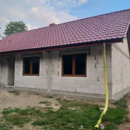 Niedokończony budynek jednorodzinny z czerwonym dachem, widoczne okna z brązowymi ramami, ściany z białych bloczk&oacute;w, piasek i gruz przed budynkiem, ż&oacute;łty wąż wentylacyjny przy elewacji.