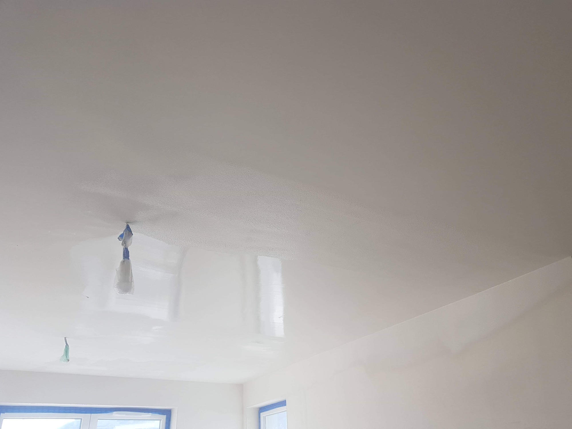 Biały sufit po gładzi, zabezpieczone taśmą okna i lampy wiszące w folii, widoczne niedoskonałości powierzchni przed malowaniem.