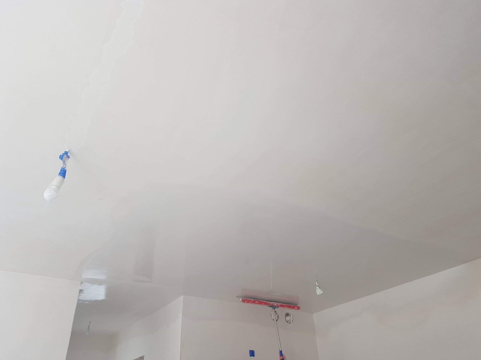 Gładka, biała powierzchnia sufitu po nałożeniu masy, widoczne narzędzie - szpachla z czerwoną gumą na teleskopowym kiju, zabezpieczone lampy i narożniki taśmą malarską.
