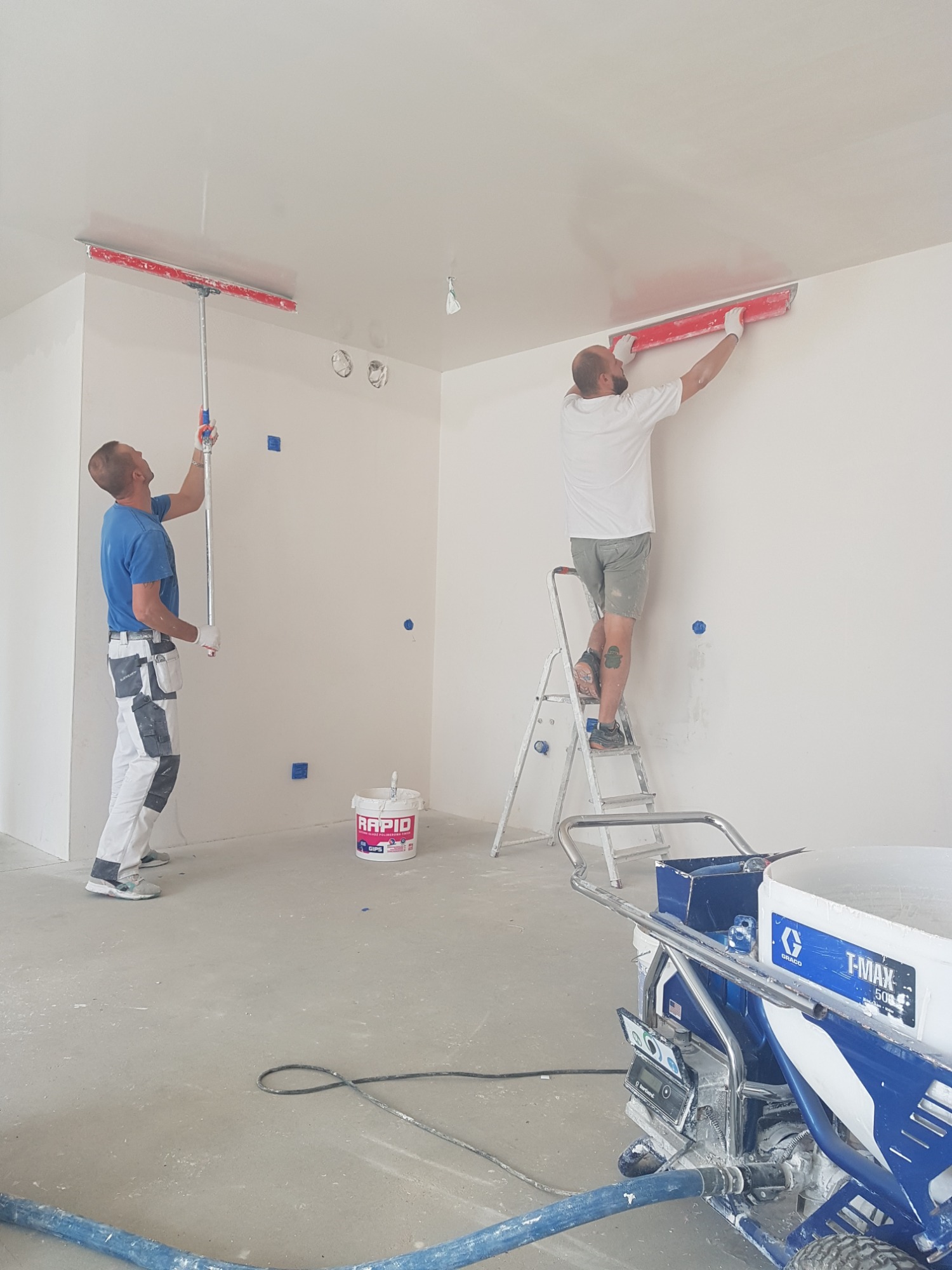 Dwóch mężczyzn wygładza sufit i ścianę w jasnym pomieszczeniu za pomocą długich szpachelek, widoczny agregat malarski Graco T-Max 506, niebieskie kwadraty maskujące na ścianie.