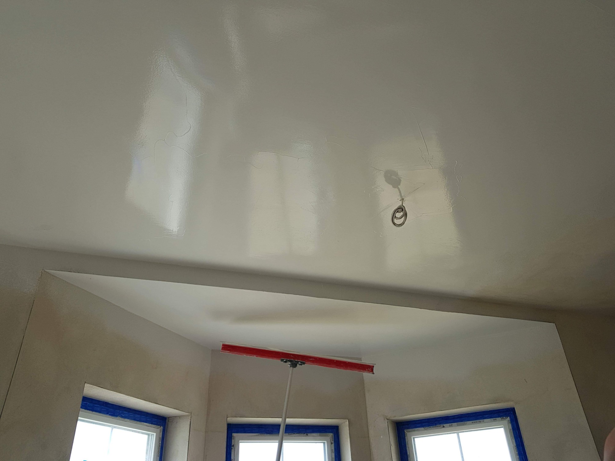 Świeżo wygładzona powierzchnia sufitu z widocznymi śladami po szpachli, uchwyt na żarówkę na środku, ściany przygotowane do malowania, okna zabezpieczone taśmą malarską.