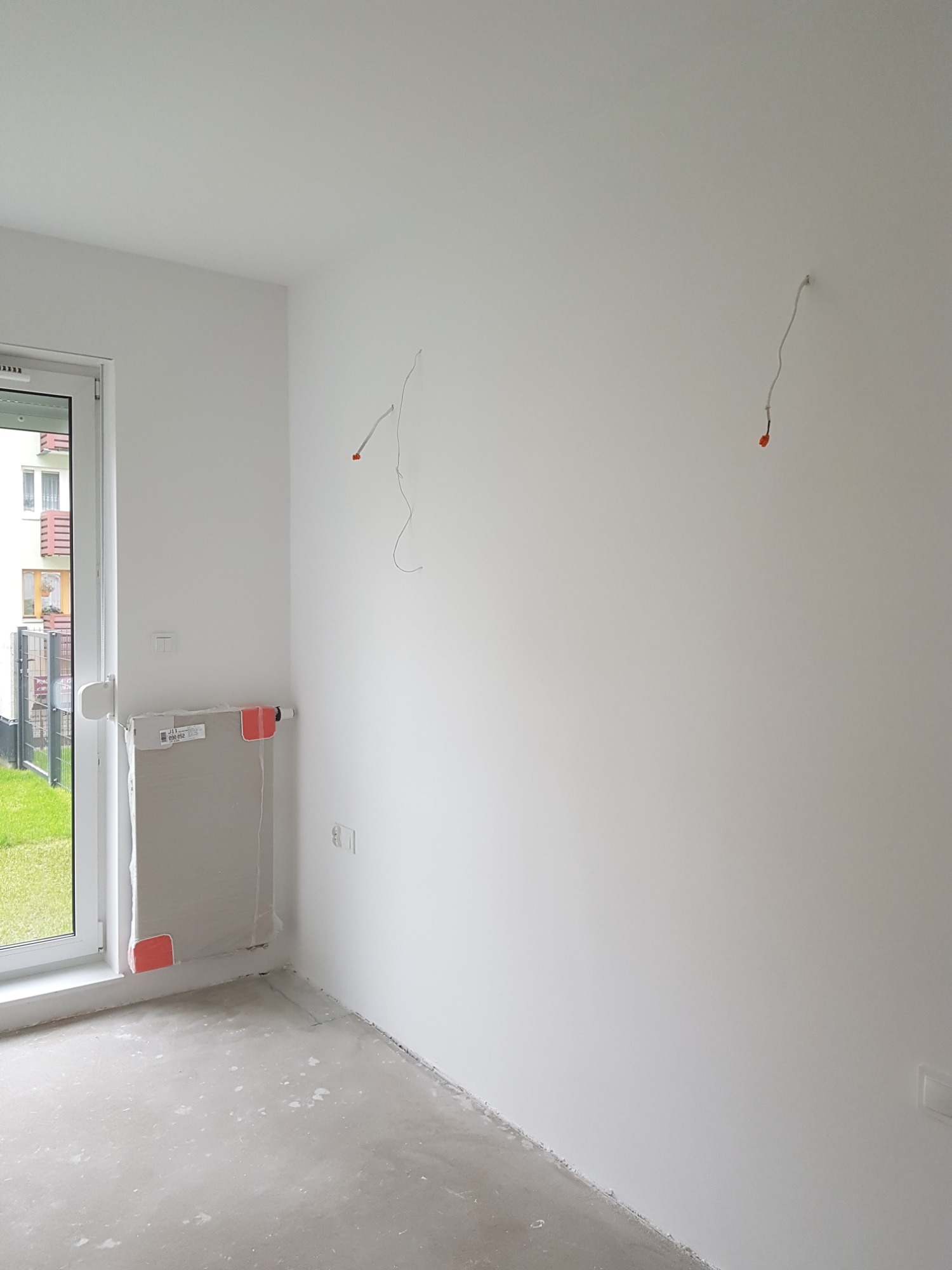 Wnętrze pokoju w trakcie szpachlowania ścian, zabezpieczony grzejnik pod oknem folią, wystające przewody elektryczne ze ściany.