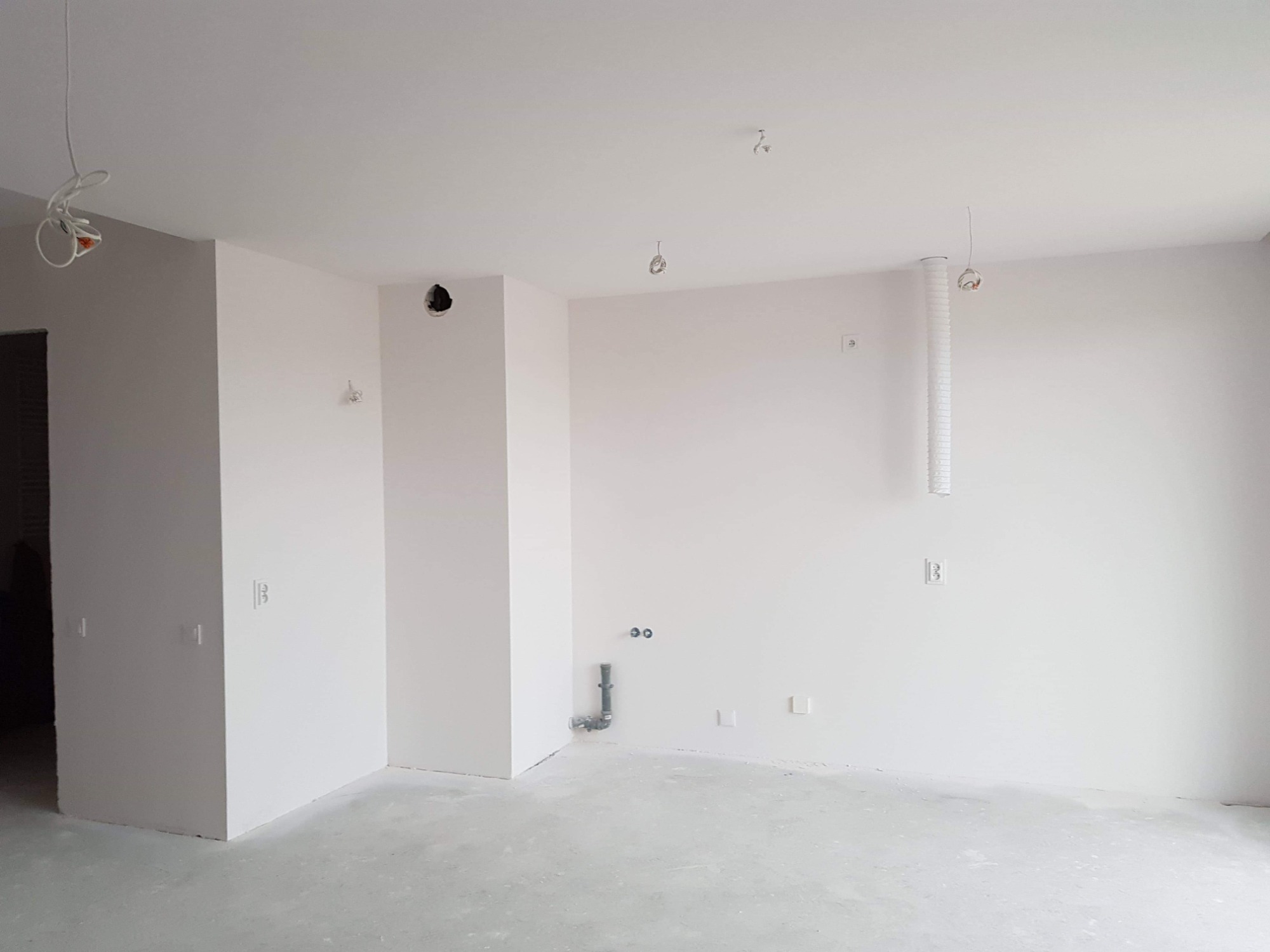 Wnętrze pokoju po malowaniu ścian i sufitu na biało, widoczne wystające kable elektryczne, rury i gniazdka elektryczne przed montażem osprzętu.