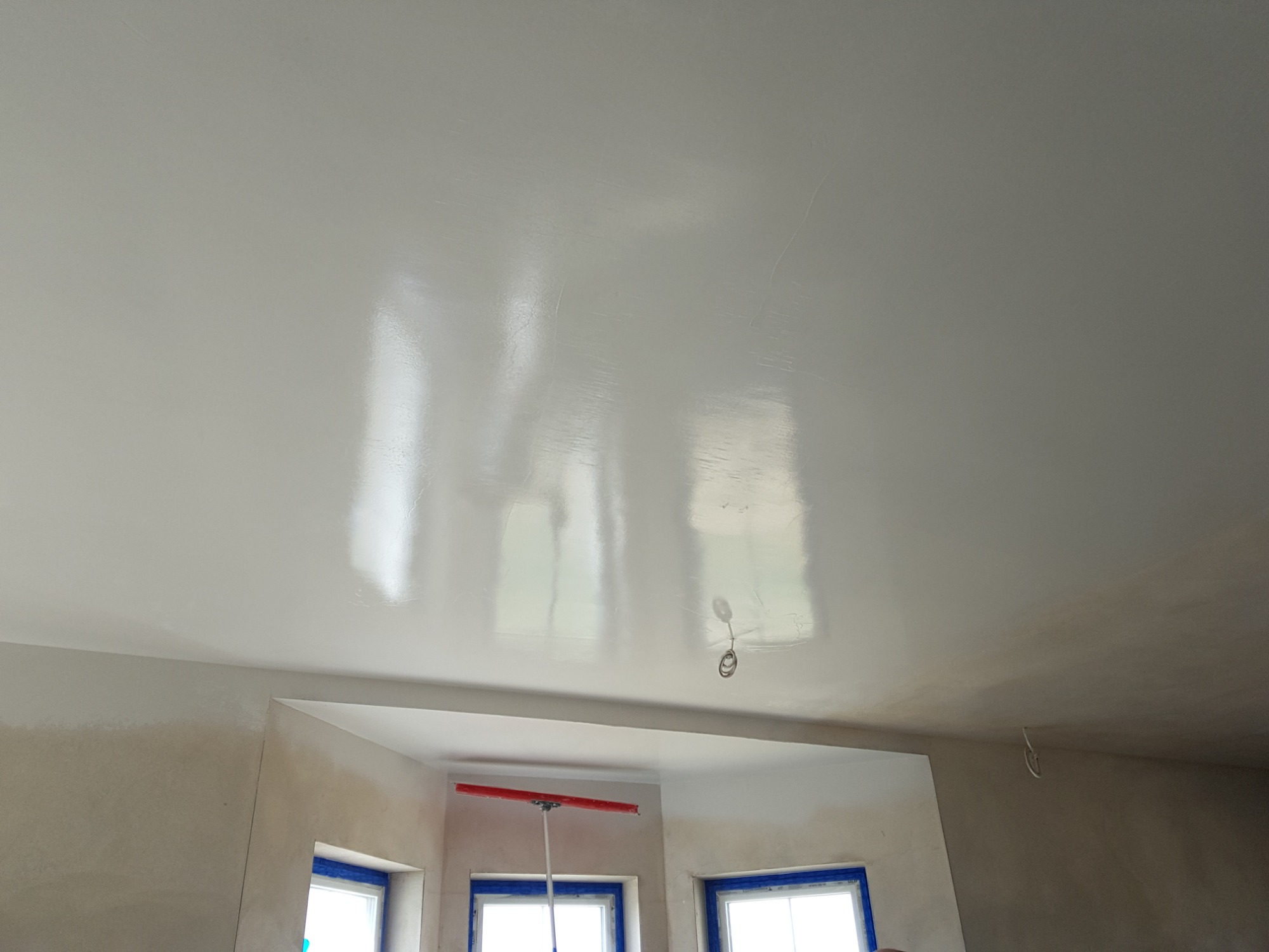 Gładka, świeżo położona biała gładź na suficie, z widocznymi refleksami światła, okna zabezpieczone niebieską taśmą malarską, w tle widoczny szpachel z długim uchwytem.