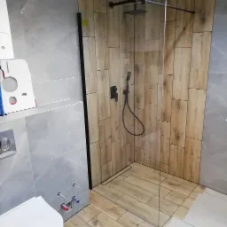 Nowoczesna łazienka z kabiną prysznicową typu walk-in, wykończona płytkami imitującymi drewno i szarym marmurem, z czarną armaturą i odpływem liniowym.