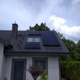 Panele słoneczne na dachu domu jednorodzinnego z ciemną dachówką, widoczne okna dachowe, komin i fragment ogrodu z roślinnością.