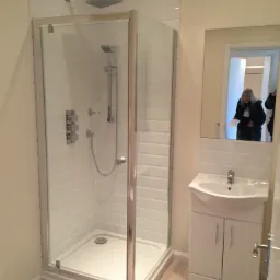 Nowoczesna kabina prysznicowa z deszczownicą i panelem sterowania, umywalka z szafką w wyremontowanej łazience, widoczne odbicie osoby w lustrze.