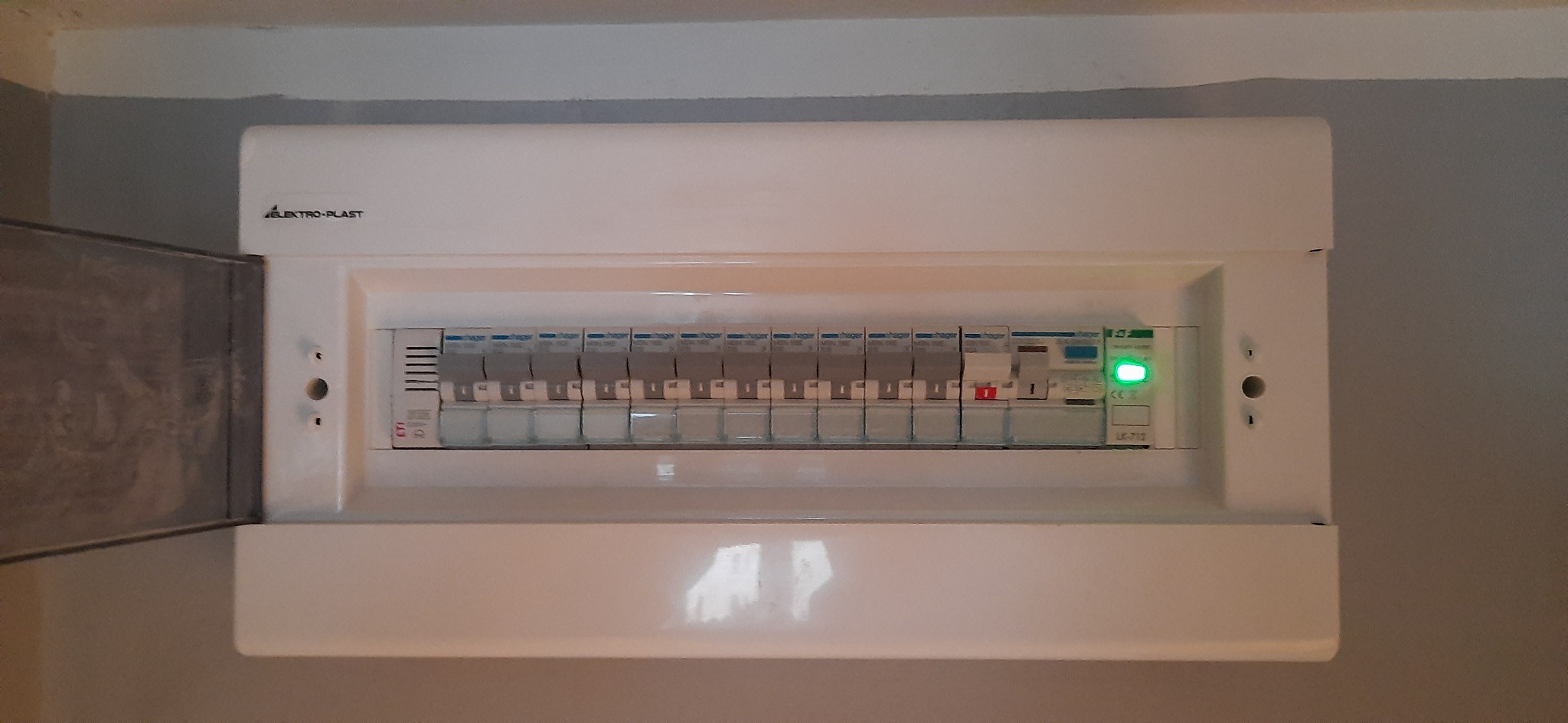 Otwarta skrzynka elektryczna Elektro-Plast z wyłącznikami Hager, jeden wyłącznik z czerwoną dźwignią, jeden z zieloną diodą, zamontowana na szarej ścianie.