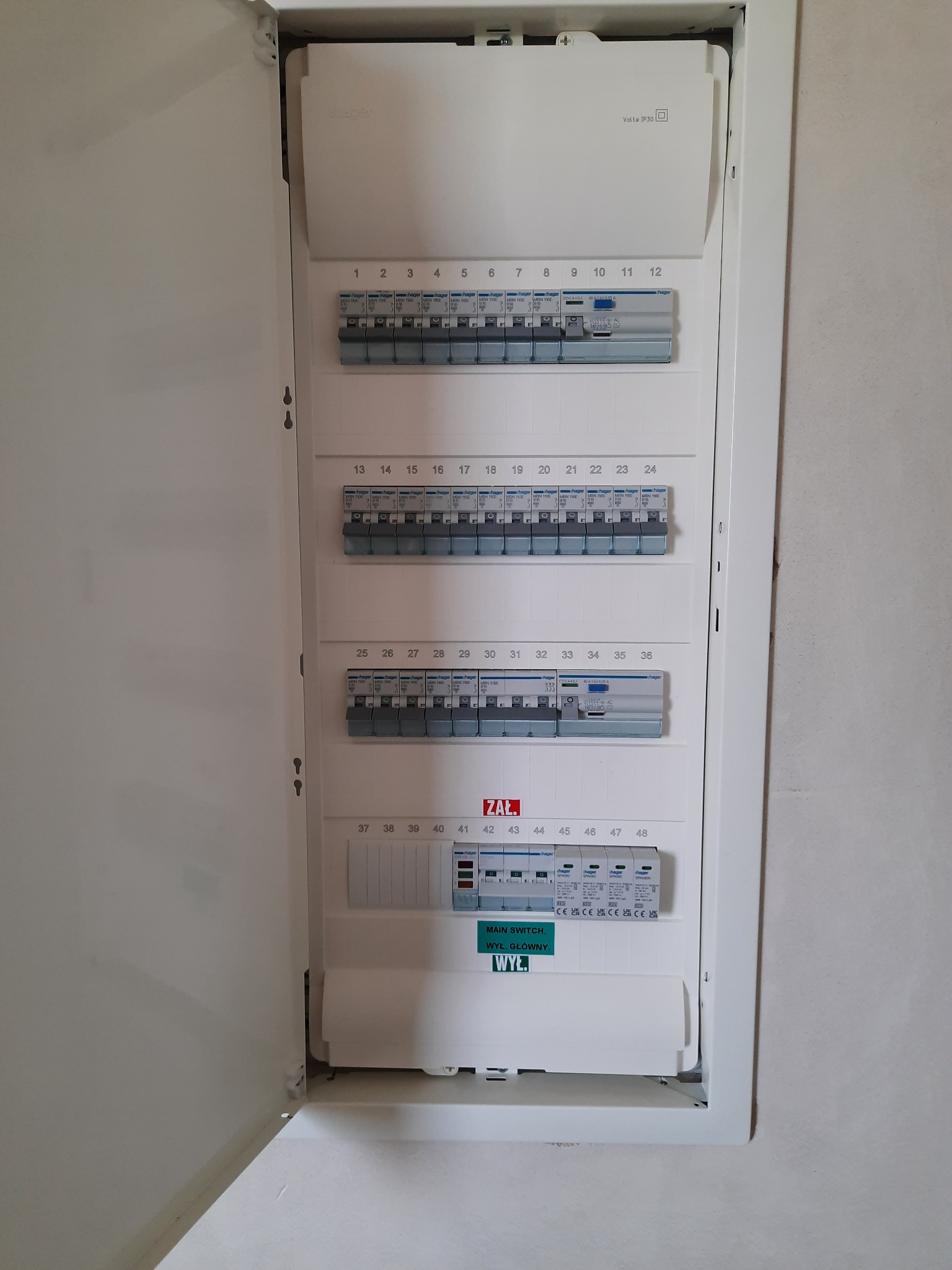 Otwarta skrzynka elektryczna z widocznymi bezpiecznikami i wyłącznikami, zamontowana na ścianie. Widoczna naklejka z napisem 'MAIN SWITCH. WYŁ. GŁÓWNY. WYŁ.'