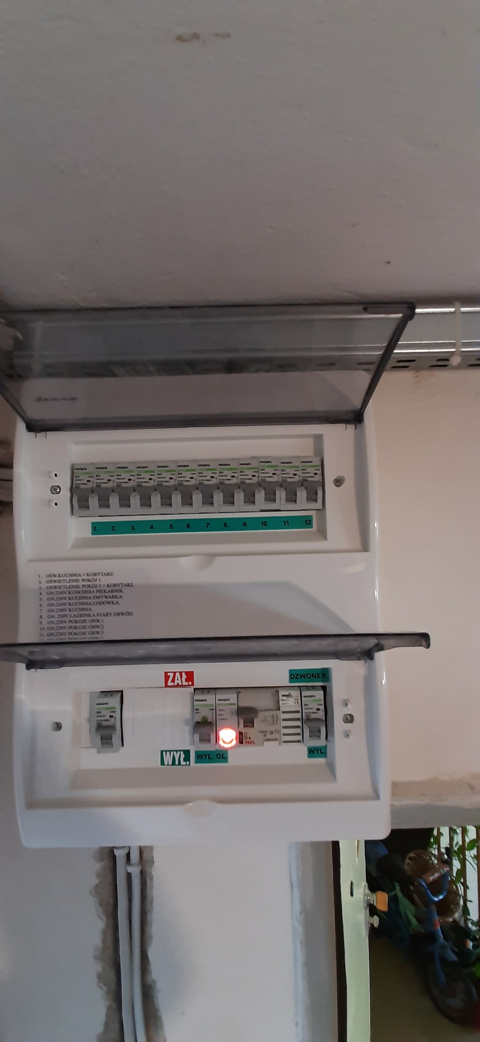 Otwarta skrzynka bezpiecznikowa z opisami obwodów, widoczne wyłączniki, jeden z nich z czerwoną diodą sygnalizującą wyłączenie, szare kable w ścianie poniżej.