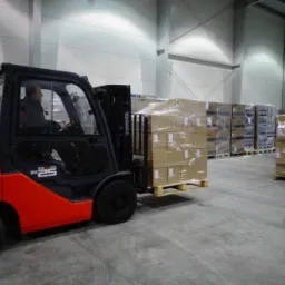 Wnętrze magazynu z wózkiem widłowym Toyota SAS 25 transportującym paletę z zapakowanymi kartonami, w tle regały z towarem oraz pracownik z wózkiem paletowym.