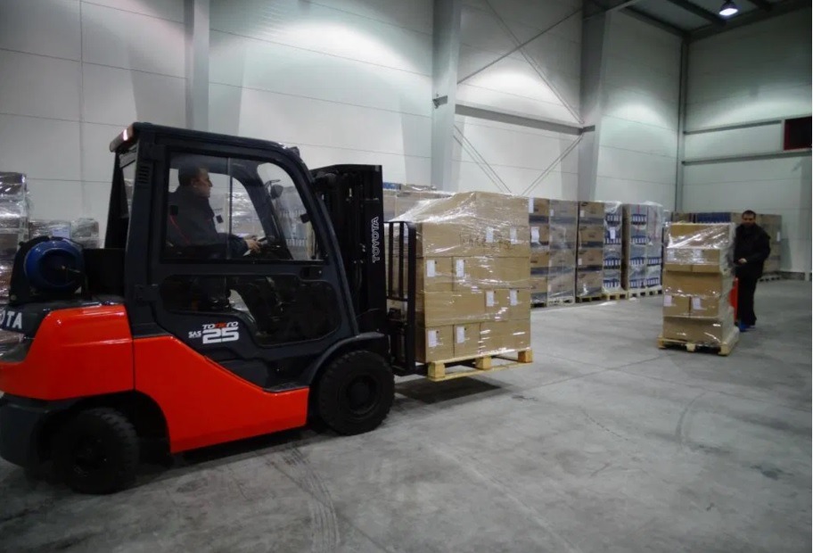 Wnętrze magazynu z wózkiem widłowym Toyota SAS 25 transportującym paletę z zapakowanymi kartonami, w tle regały z towarem oraz pracownik z wózkiem paletowym.