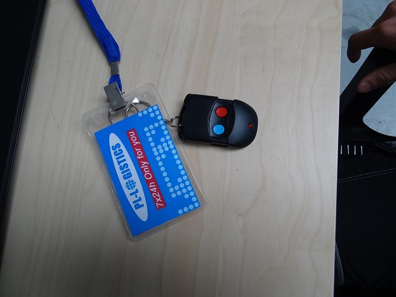 Identyfikator PL-I GISTICS 7x24h z niebieską smyczą leżący obok pilota do bramy na jasnym biurku.