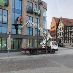 Dwóch pracowników w pomarańczowych kamizelkach roboczych na platformie podczas montażu okien w nowoczesnym budynku hotelowym z cegły, widoczne detale konstrukcyjne i tabliczka z nazwą Hotel Sova,...