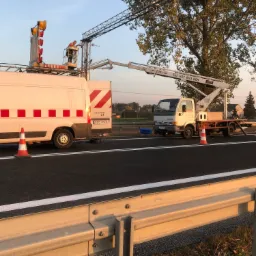 Pracownik w pomarańczowej kamizelce na platformie roboczej na wysokości, przy montażu lub konserwacji oświetlenia drogowego, z użyciem dwóch pojazdów z wysięgnikami na tle drzew i błękitnego nieba.