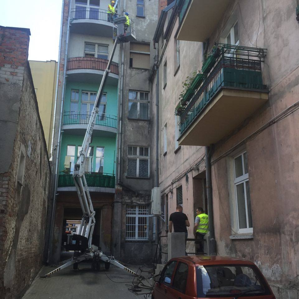 Pracownicy w koszu na wysokościach podczas prac remontowych na tle starych kamienic, widoczny samochód osobowy i wąska ulica.