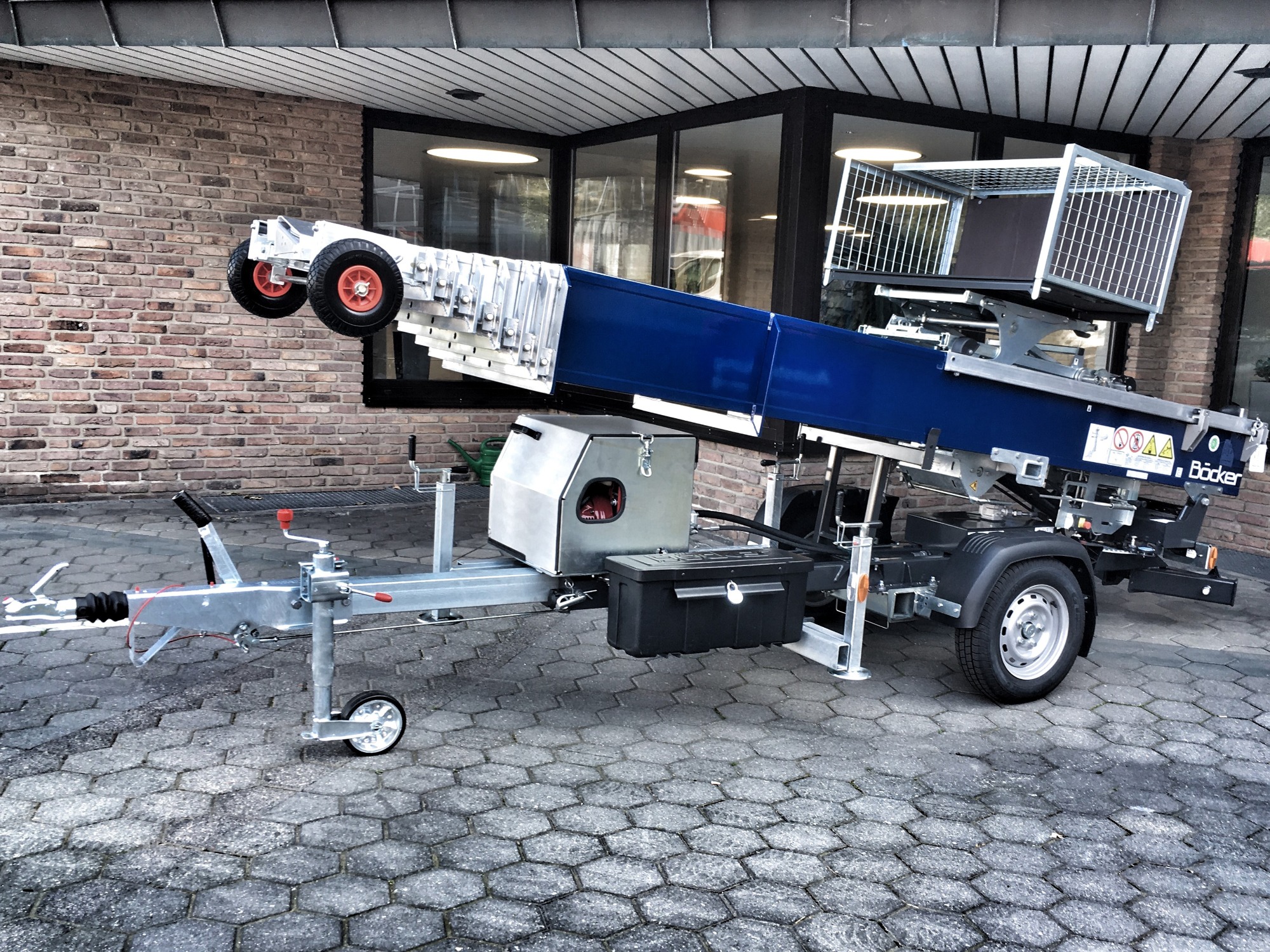 Przyczepa z platformą transportową na podwoziu teleskopowym marki Böcker ustawiona na kostce brukowej przed budynkiem z cegły i dużymi oknami.