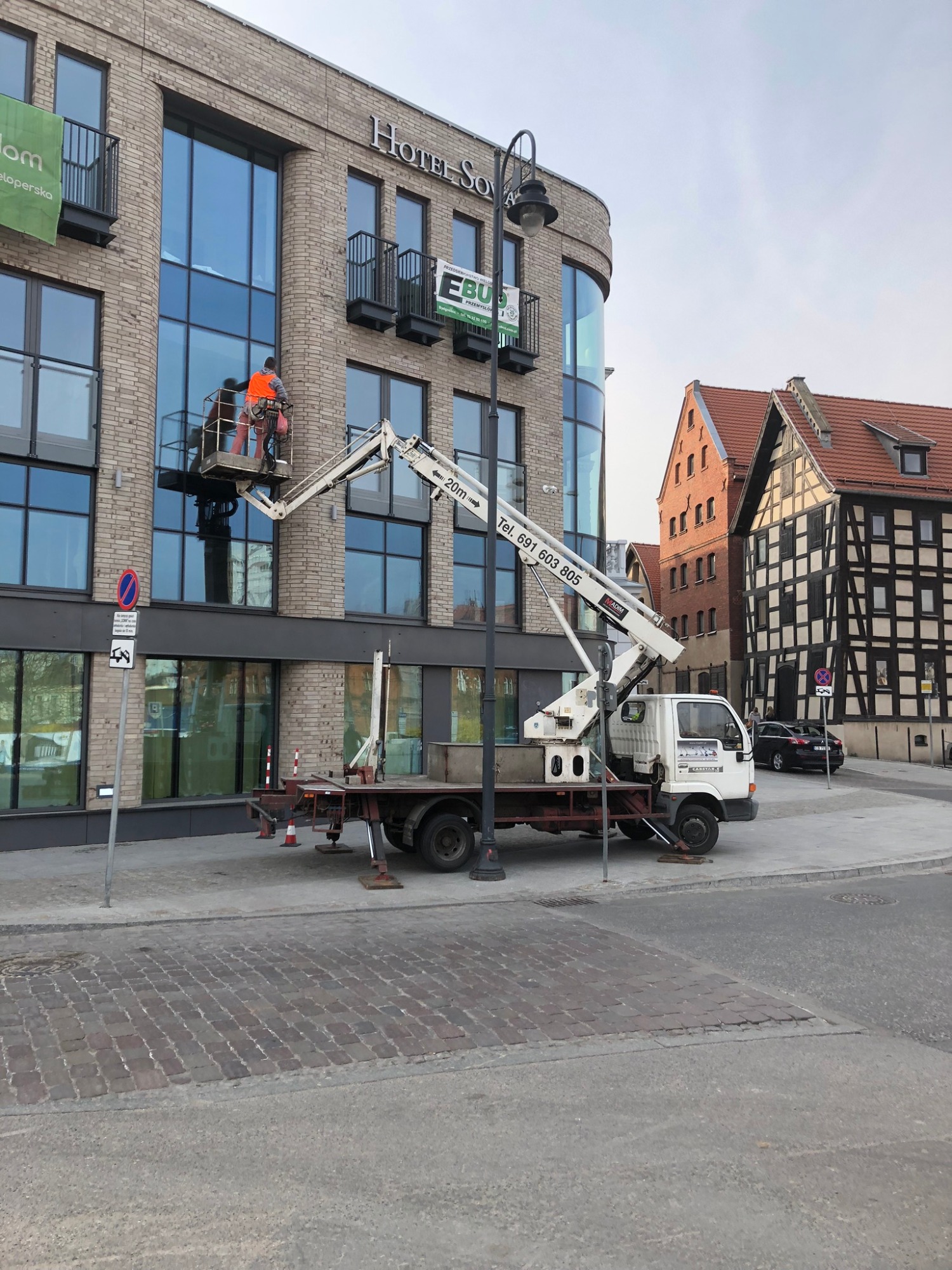 Dwóch pracowników w pomarańczowych kamizelkach roboczych na platformie podczas montażu okien w nowoczesnym budynku hotelowym z cegły, widoczne detale konstrukcyjne i tabliczka z nazwą Hotel Sova,...