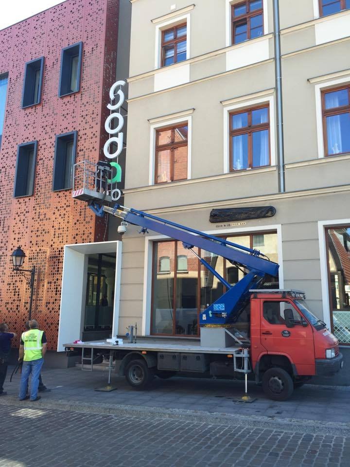 Czerwony samochód z niebieskim wysięgnikiem ustawia biały neon na elewacji budynku o dwóch różnych stylach architektonicznych.