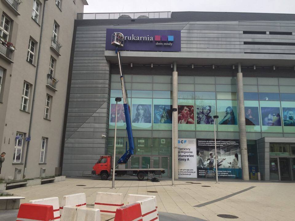 Czerwony samochód z niebieskim ramieniem podnośnika koszowego na tle szarego budynku z dużym szyldem 'Drukarnia dom mody'.