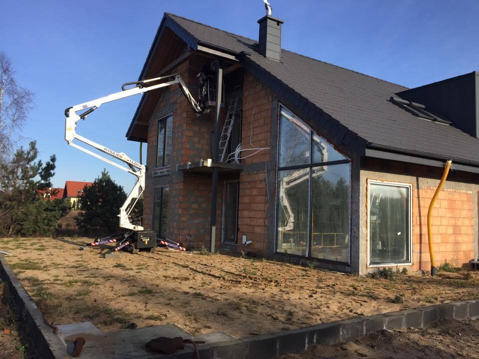 Biały podnośnik koszowy przy dwukondygnacyjnym domu w trakcie budowy, z widoczną elewacją z czerwonej cegły i dużymi oknami.