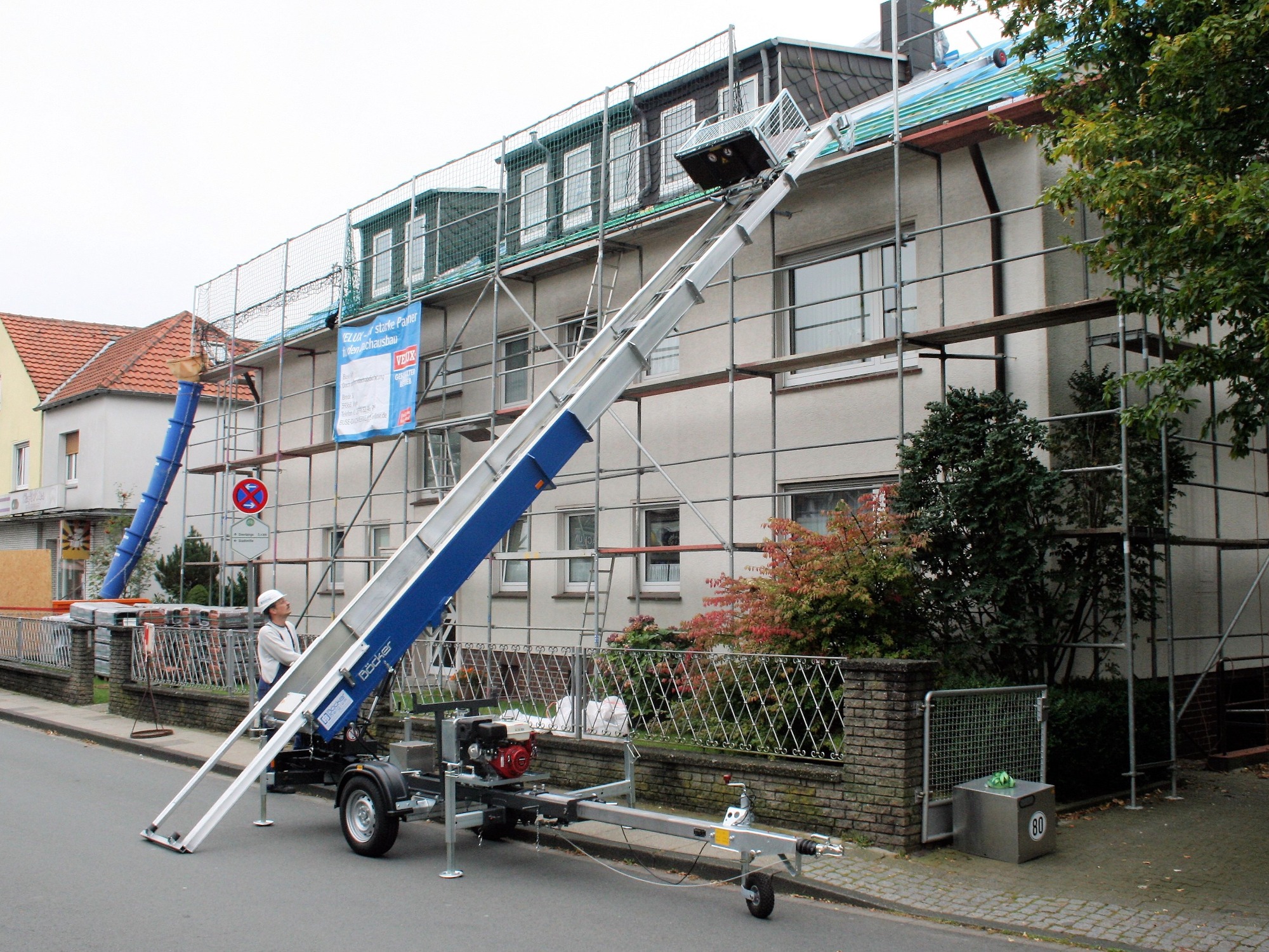 Podnośnik meblowy Böcker ustawiony przed budynkiem w rusztowaniach, pracownik w kasku kontroluje proces wnoszenia materiałów na dach.