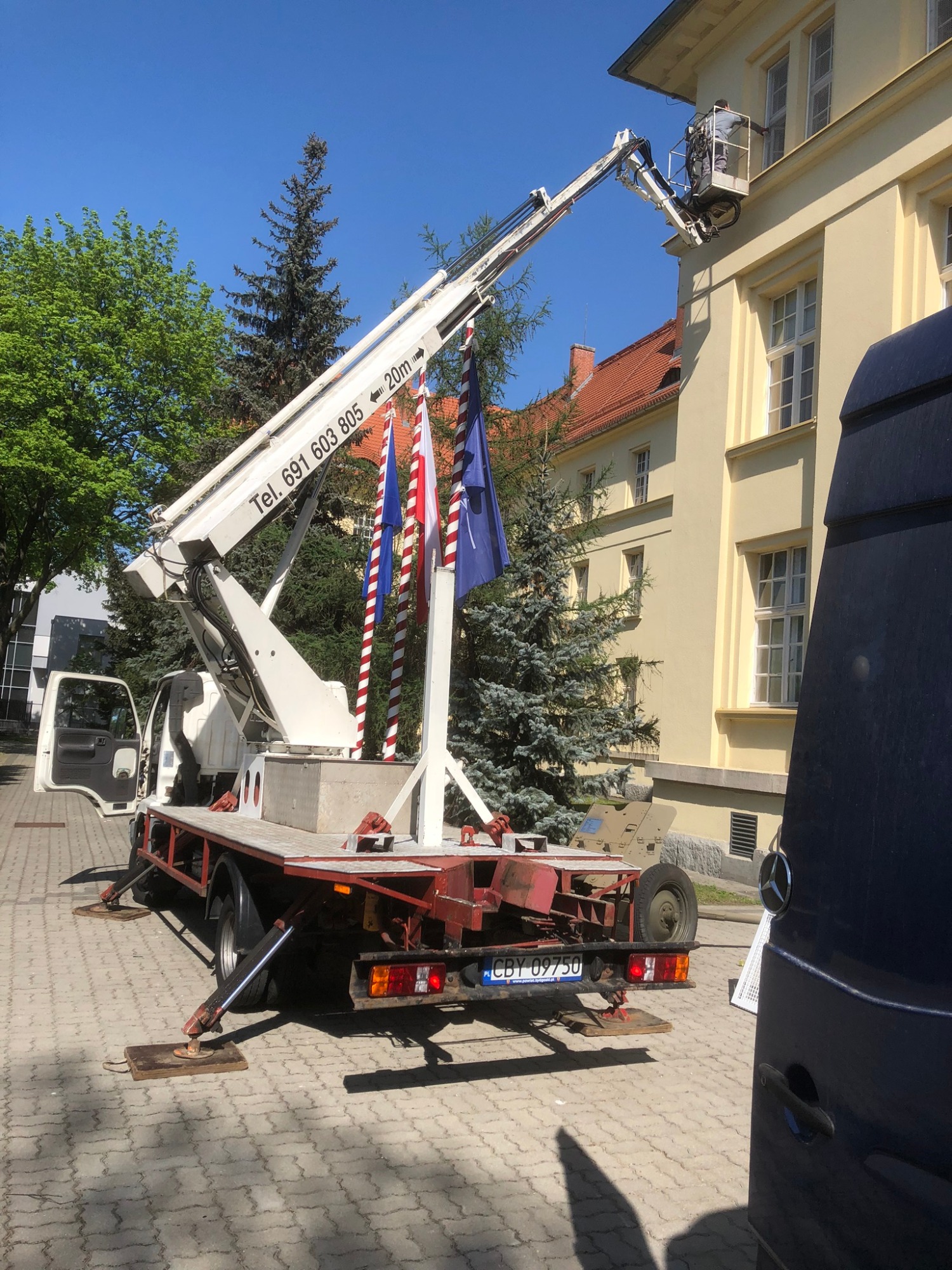 Pracownik w koszu podnośnika na wysięgniku, wykonujący prace na elewacji budynku z jasną fasadą, widoczne flagi dekoracyjne i zaparkowany samochód dostawczy.