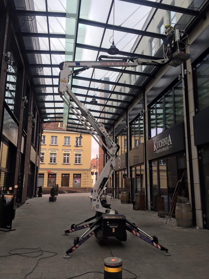 Pracownik w koszu podnośnika teleskopowego, konserwujący szklany dach pasażu handlowego, widoczne budynki w tle.