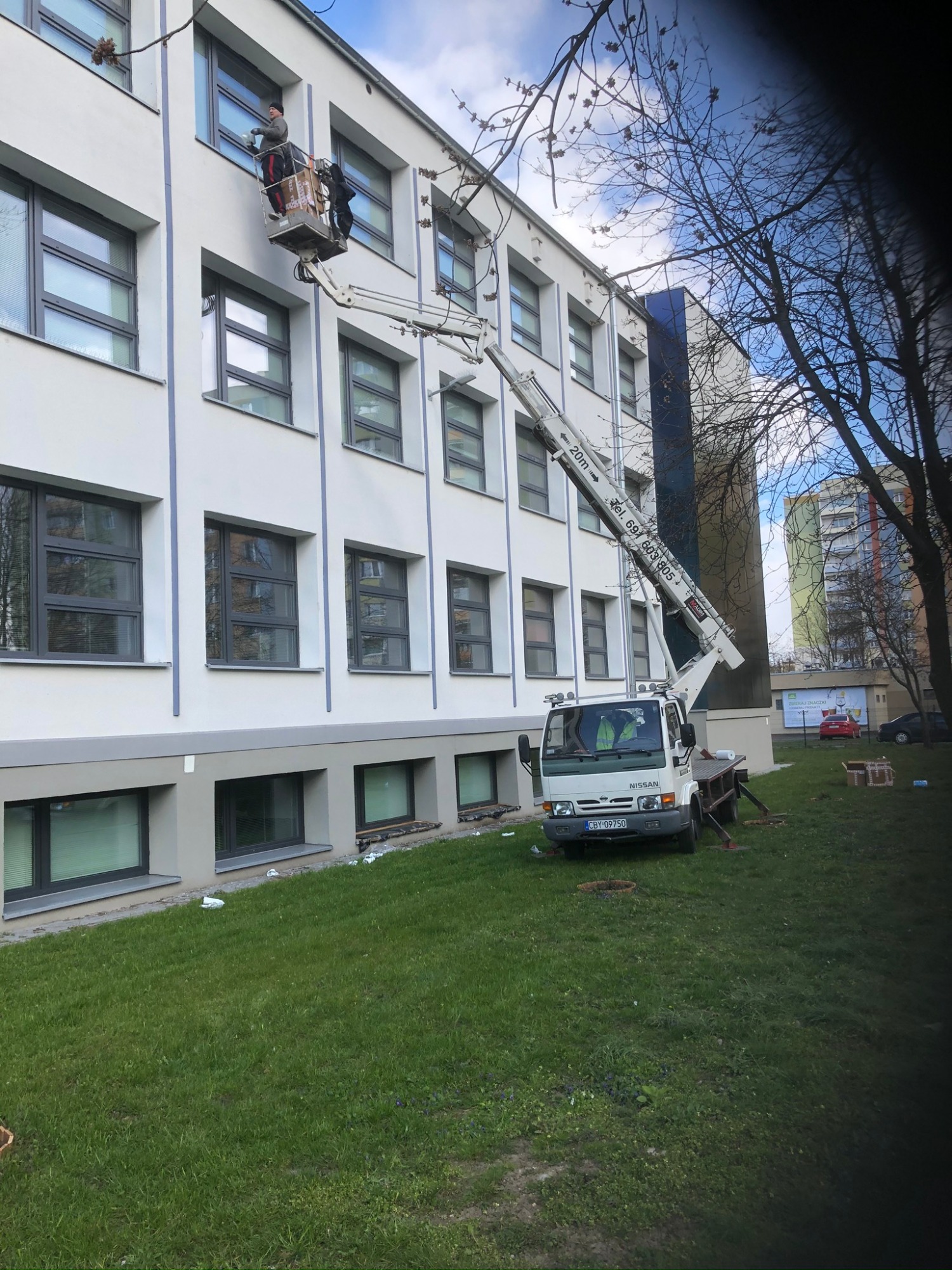 Pracownik w koszu białego pojazdu z wysięgnikiem, czyści okna na białym budynku z szarymi ramami okiennymi, na trawniku przed budynkiem