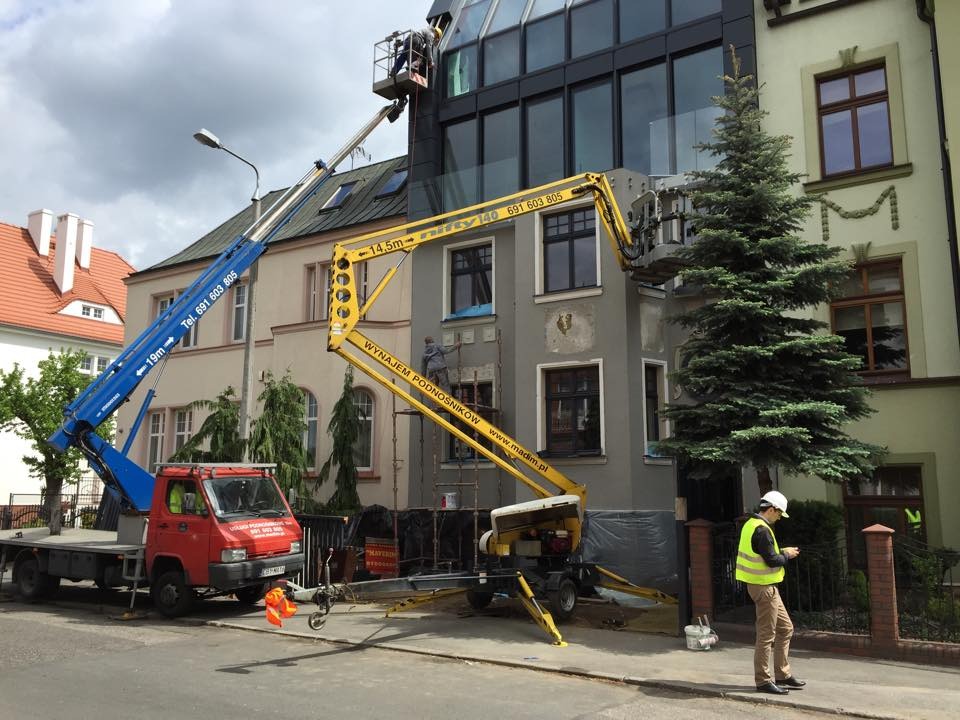 Dwa podnośniki teleskopowe, niebieski i żółty, używane do prac elewacyjnych na budynku w miejskiej scenerii. Pracownicy w koszach wykonują prace na różnych wysokościach. Na pierwszym planie...