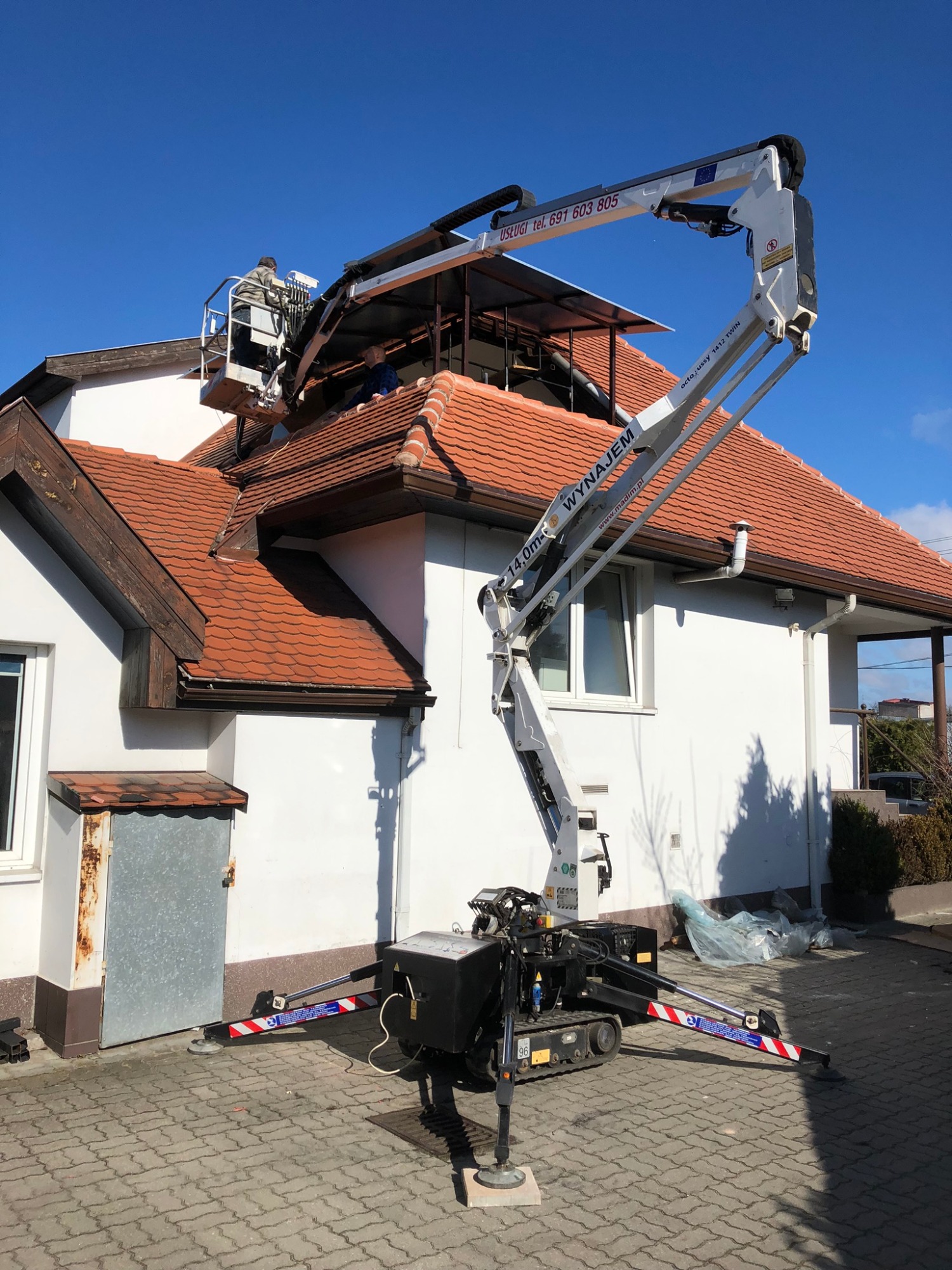 Prace na dachu domu jednorodzinnego z użyciem samojezdnego urządzenia z wysięgnikiem teleskopowym, dwuosobowa platforma robocza, zabezpieczone podpory stabilizujące, błękitne niebo.