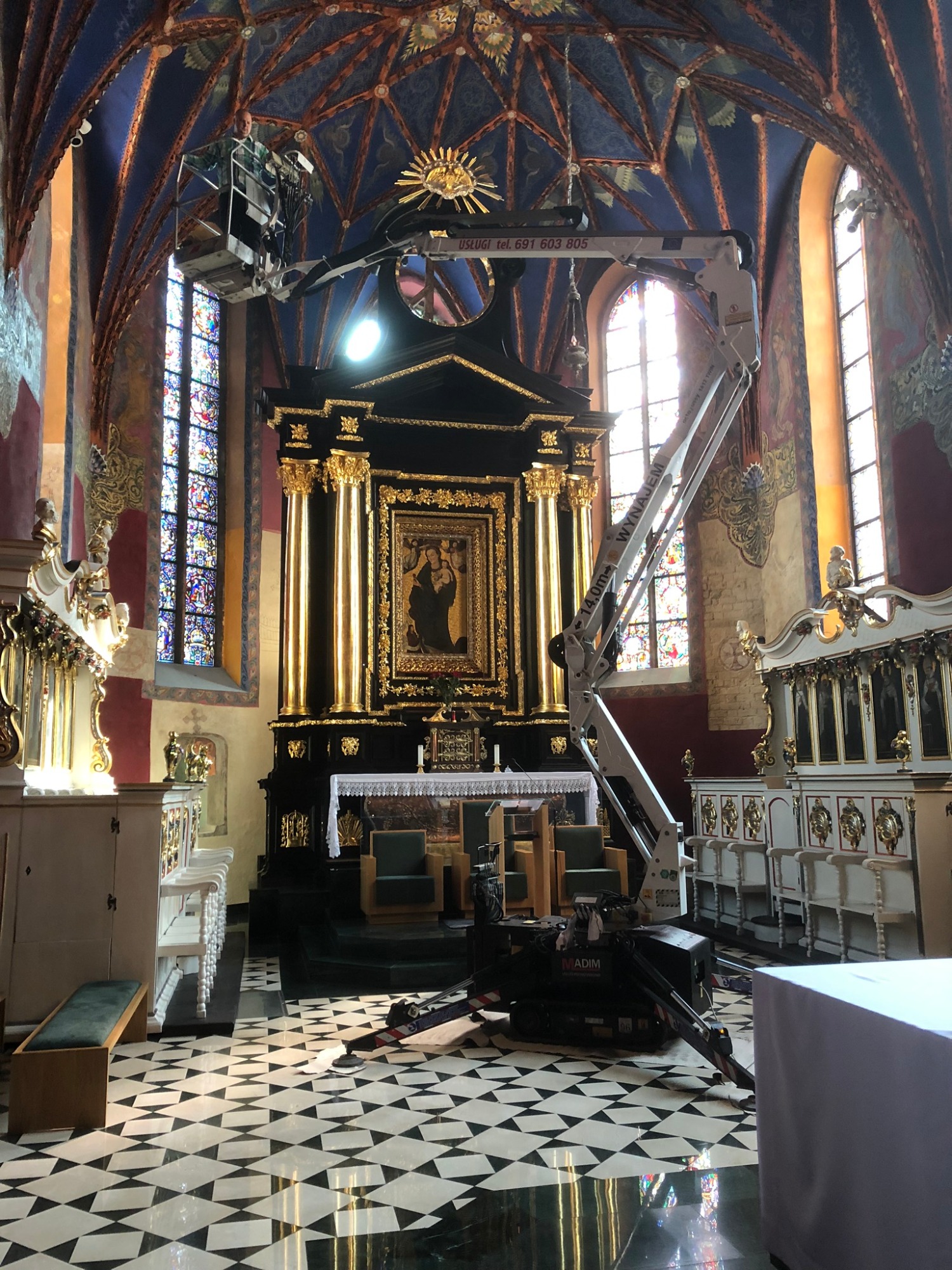 Wnętrze kościoła z czarno-białą podłogą, gdzie pracownik w koszu podnośnika dokonuje prac konserwatorskich przy ołtarzu głównym, widoczne witraże w oknach.